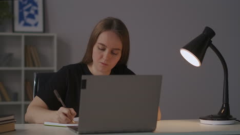 e-aprendizaje en casa estudiante está viendo la conferencia en línea y escribiendo notas en el cuaderno retrato de la mujer en la mesa por la noche la lámpara de la mesa está encendiendo