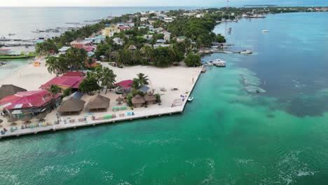 vista de avión no tripulado en belice volando sobre el mar verde y azul del caribe, un cay de arena blanca con palmeras y restaurantes con un barco cruzando un canal en un día nublado