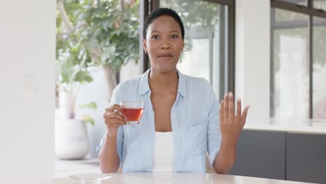 Fröhliche-Afroamerikanische-Frau,-Die-Am-Tisch-Sitzt,-Tee-Trinkt-Und-Einen-Videoanruf-Führt,-Zeitlupe