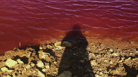 Shadow-Of-A-Person-Standing-On-Rocky-Shore-Of-A-Toxic-Mining-Lagoon-In-Twelveheads,-Cornwall,-UK