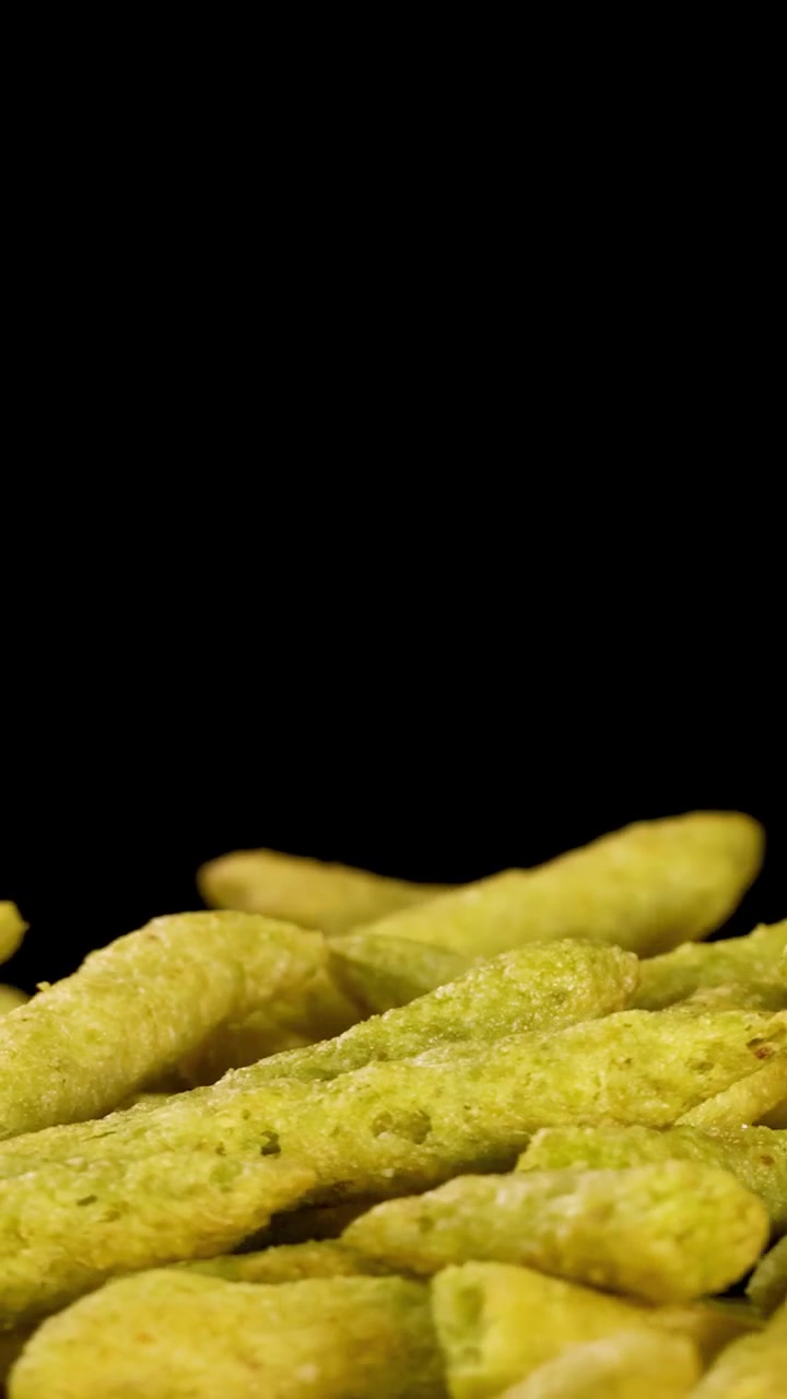 Hand lifts green pea puffed chip snack under bright studio lighting against black background.