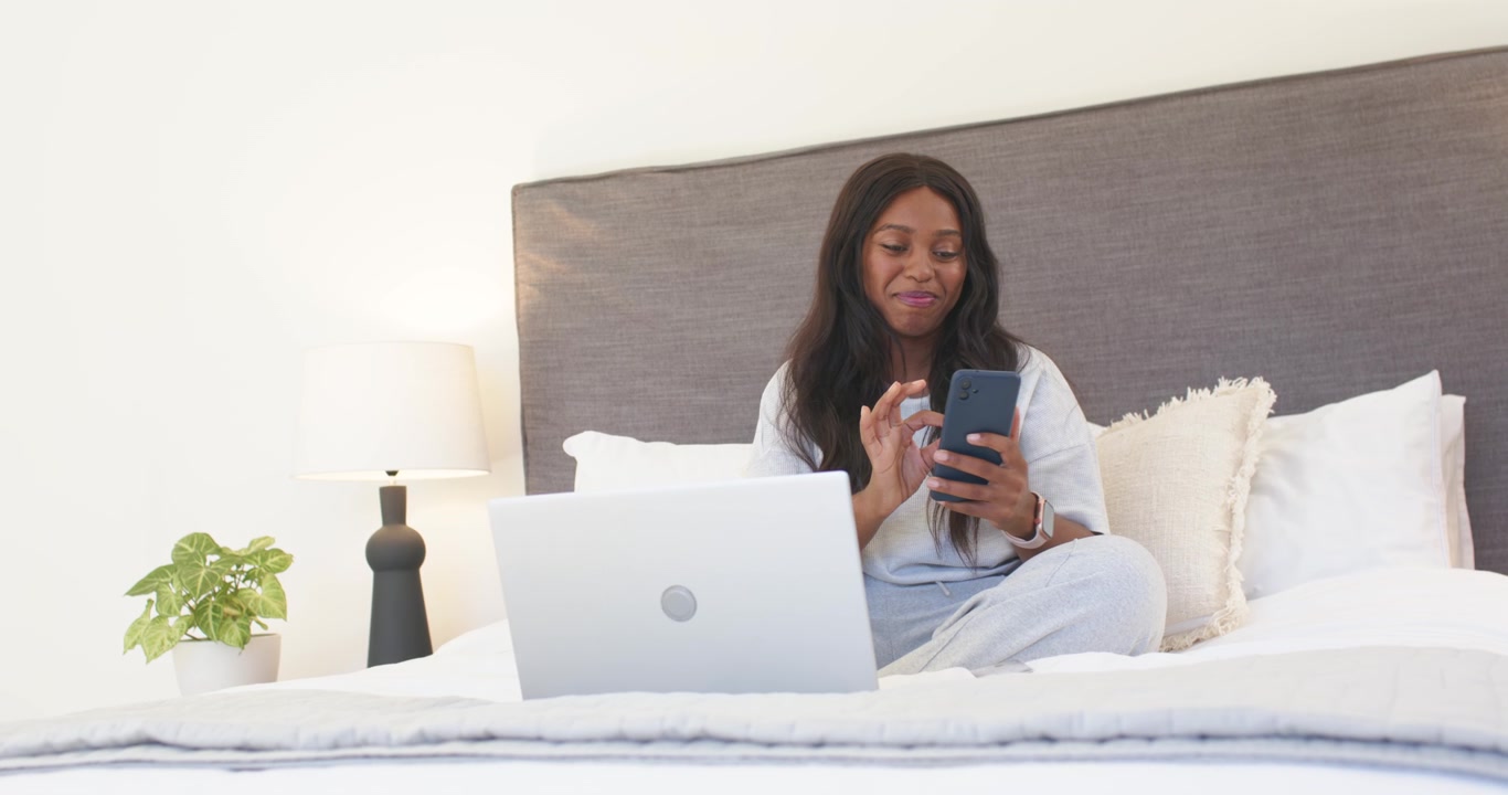 Phone pinging African woman sitting on bed, scrolling phone, checking laptop and reading messages