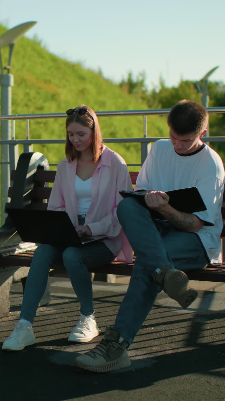 Woman in pink seated outdoors studying with friend, she withdraws hand from his tablet, continues on her laptop, with green hill and light poles in the background
