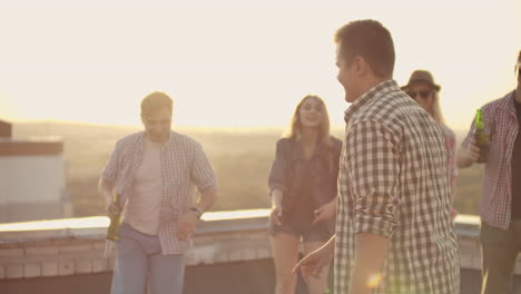 a young man in a gray plaid shirt moves in dance with his friends. he enjoys and has fun with beer on the roof.