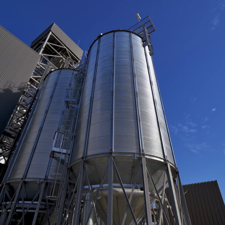 Modern silos on blue sky background. Large silver grain elevator outdoors. Industrial territory of warehouse. Agricultural storage and processing plant.