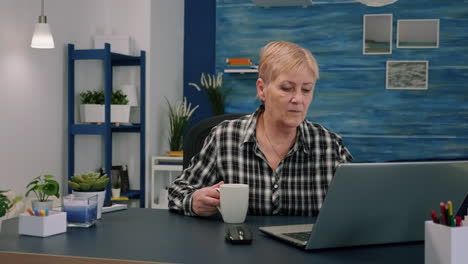 Old-businesswoman-sitting-at-workplace-typing-at-laptop,-drinking-coffee