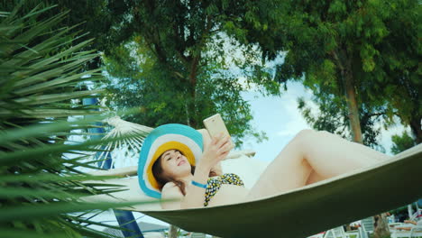 a modern young woman in a wide-brimmed hat and open swimsuit enjoys a holiday on a hammock reads new