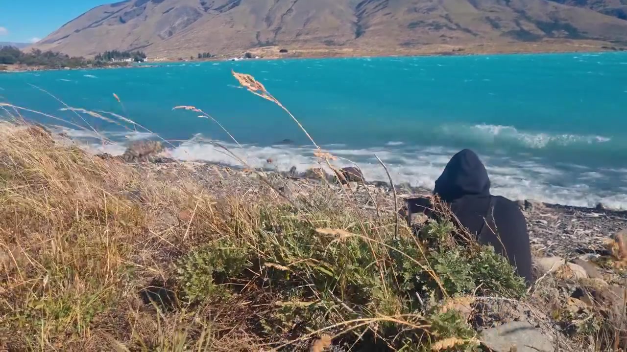 Un uomo filma le acque turchesi del Lago Argentino a Puerto Bandera in una giornata soleggiata e ventosa, catturando il remoto paesaggio patagonico e un'esperienza turistica panoramica.