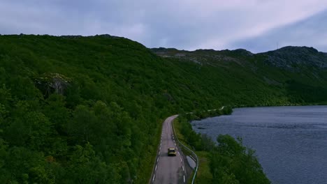 Siguiendo-Un-Auto-Dorado-A-Través-De-Los-Exuberantes-Bosques-Naturales-Verdes-Del-Norte-De-Noruega-En-Lofoten-Con-Un-Lago-Azul-A-La-Derecha-Y-Montañas-A-La-Izquierda-Y-El-Horizonte