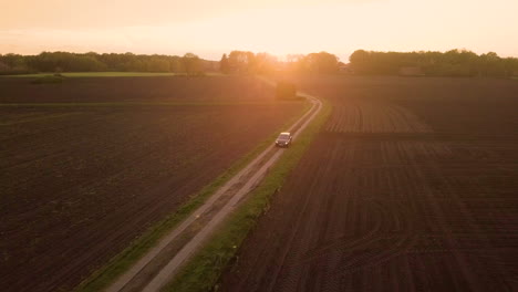 vista aérea de gran ángulo de un coche de seguimiento que atraviesa una carretera de campo vacía con tierras de cultivo vacías durante la puesta del sol y el amanecer en la región rural