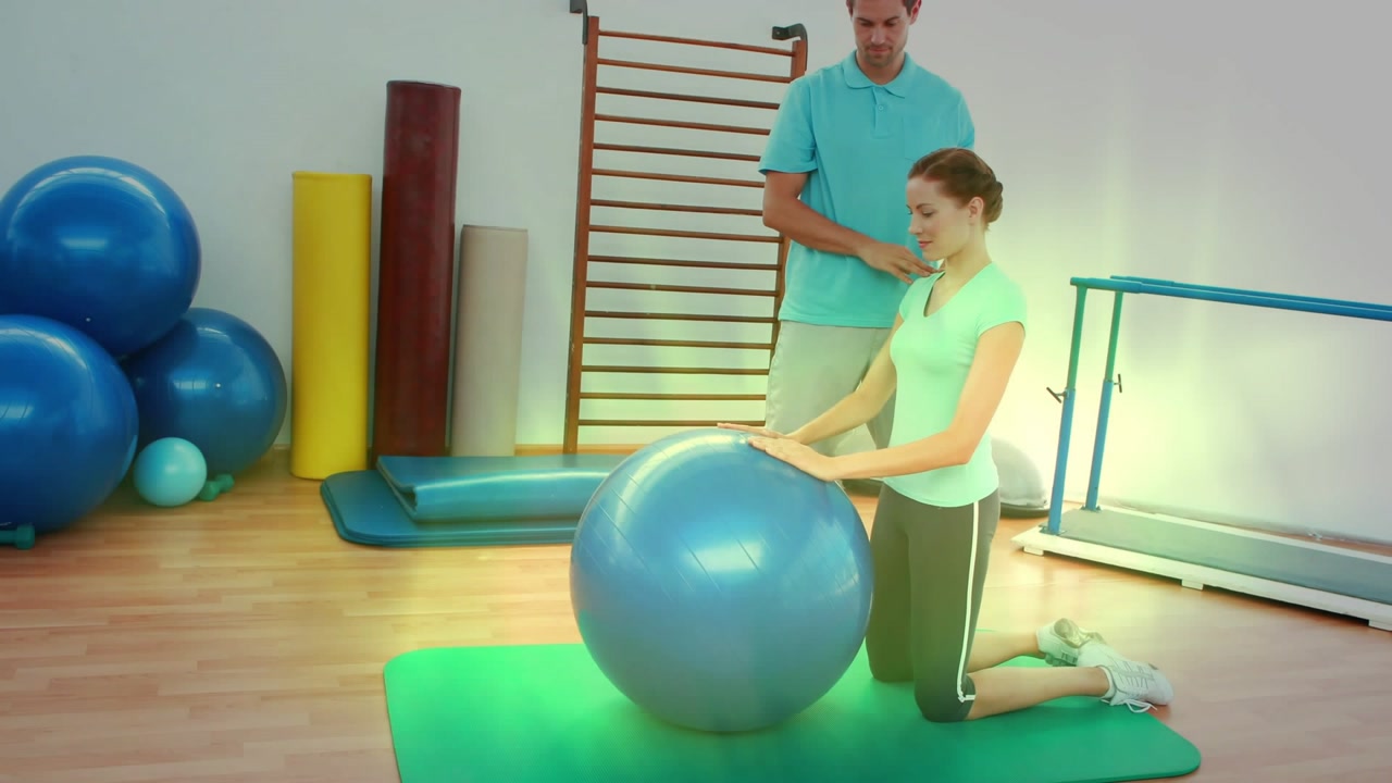 Woman kneeling on green mat placing hands on blue ball, trainer guiding shoulders for rehab stretch