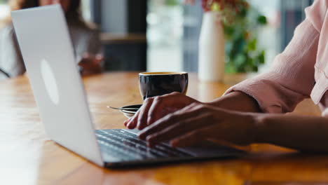 Junge-Geschäftsfrau-Mit-Kaffee,-Die-Am-Laptop-Arbeitet,-Sitzt-Im-Café-Mit-Einem-Kollegen-Im-Hintergrund