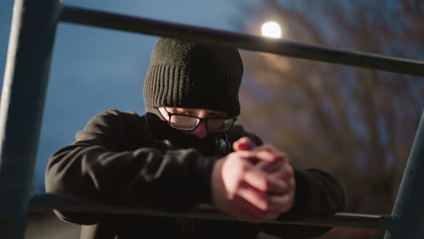 close-up of a young boy wearing glasses and a black jacket, resting on an iron bar with his head bowed, the background features blurred lights and bare trees