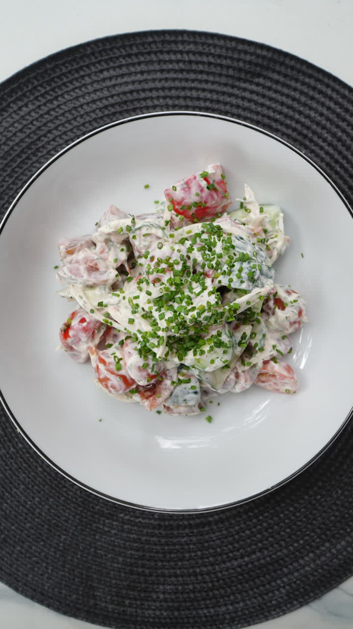 Vertical footage White plate with fresh tomato and cucumber salad with yogurt dressing, garnished with chives, being placed on a black placemat