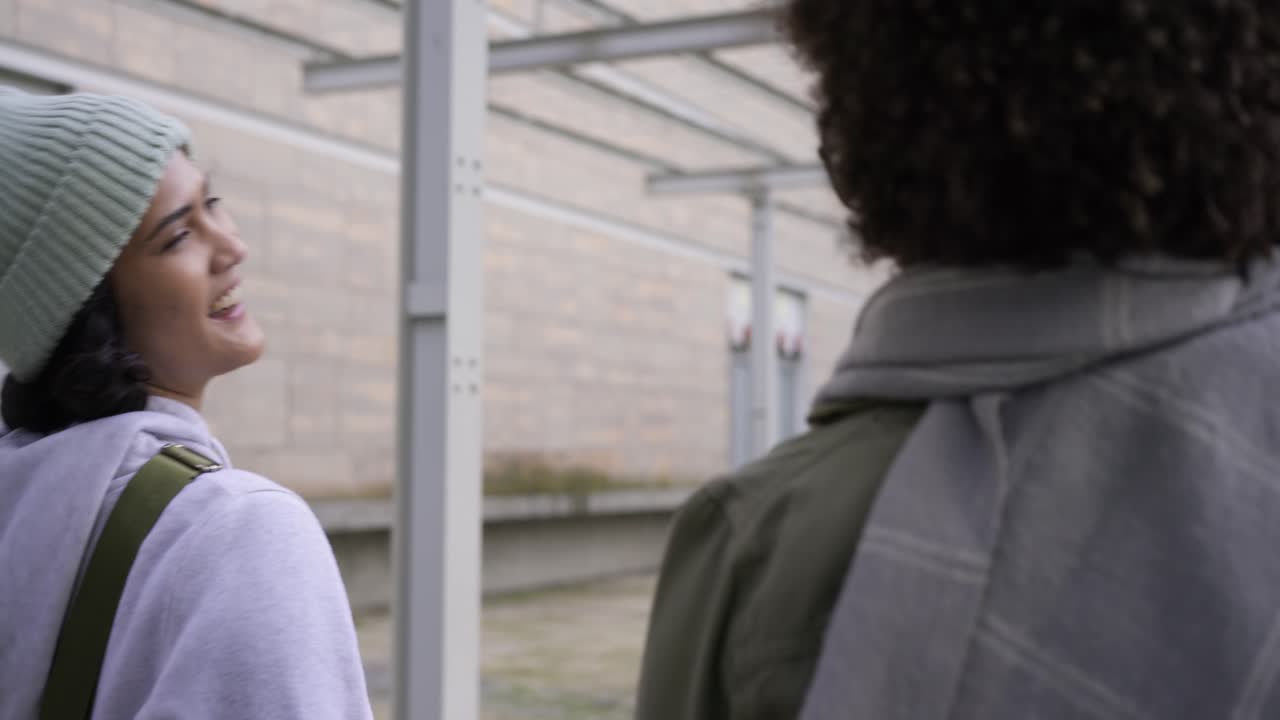 Turning head back and smiling, female friends walking under metal canopy gesturing explaining story