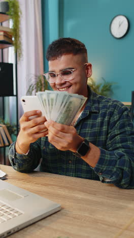 Un-Hombre-Feliz-Y-Sonriente-Contando-Dinero-En-Efectivo-En-Dólares-Usando-Ingresos-Por-Teléfono-Inteligente-Ahorra-La-Lotería-En-Casa