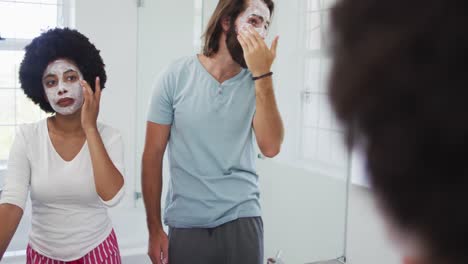 Pareja-De-Raza-Mixta-Aplicando-Mascarilla-Juntos-Mientras-Se-Miran-En-El-Espejo-Del-Baño