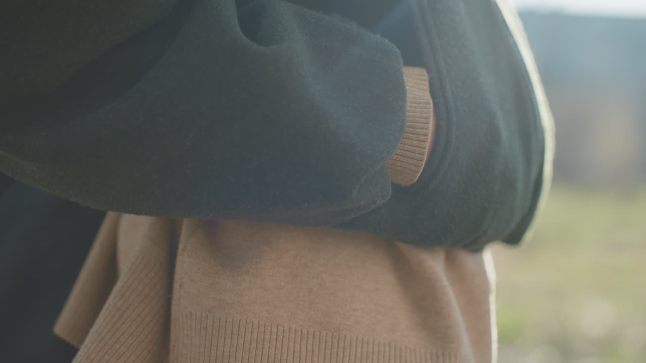 Close up of female hand with red nail polish resting on pocket of green jacket over beige sweater outdoors in natural light showing detail of casual outerwear style texture fashion