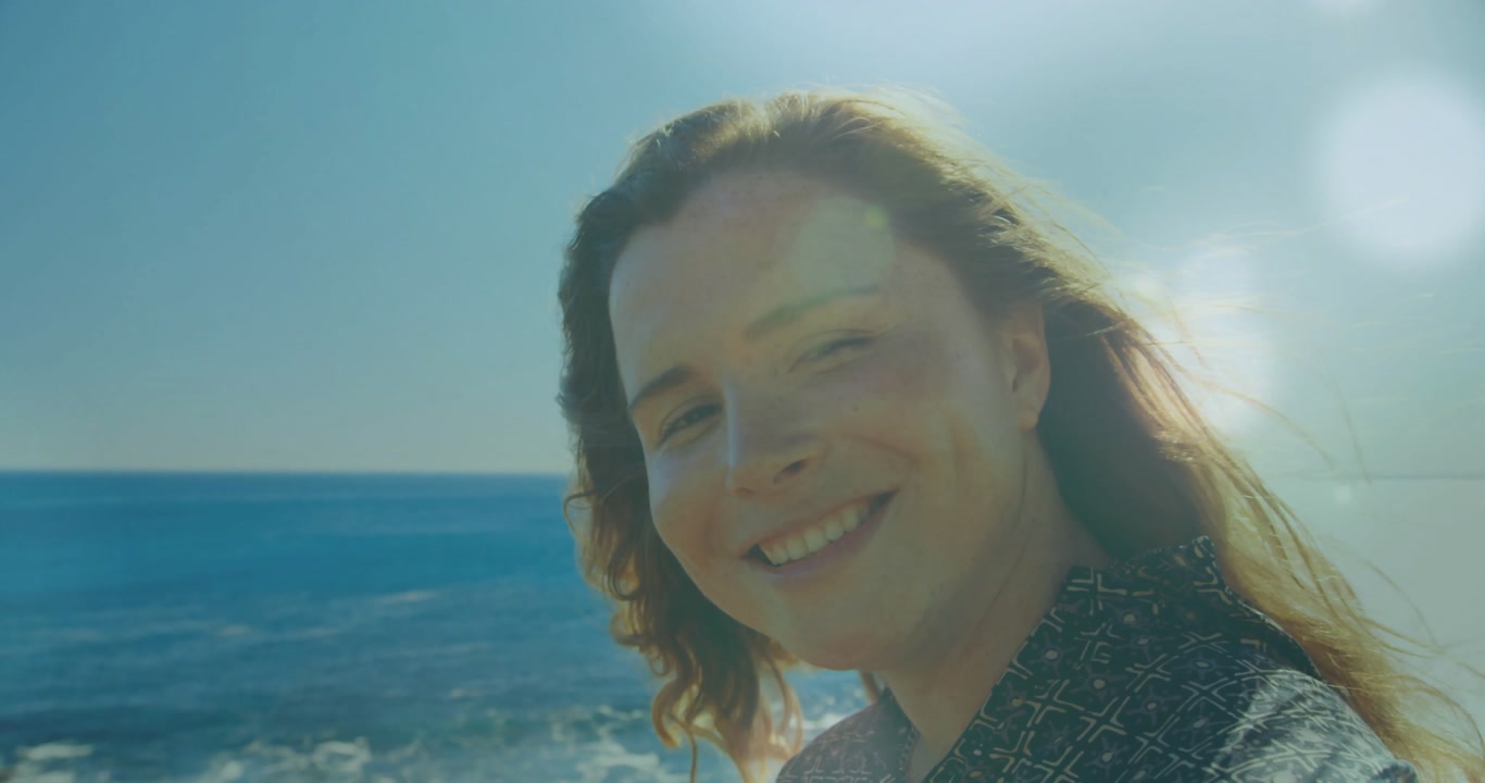 woman following sun on rocky coastal overlook, ocean breeze lifting hair, sun producing lens flares