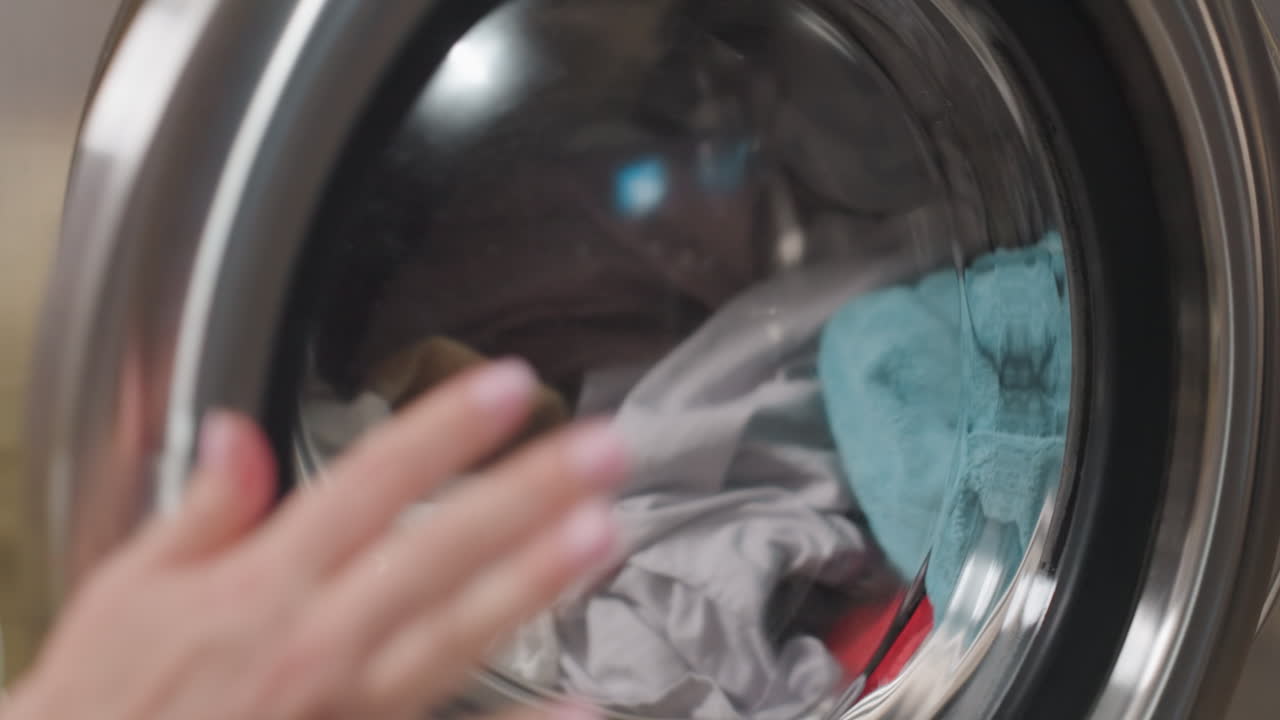 Close view shows worker loading mixed fabrics into industrial washer drum, hand pushes garments inside, gray sheet over red towel, stainless interior, smooth workflow, focus on motion and texture