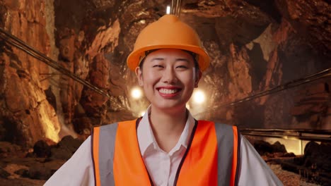 close up of asian female engineer wearing safety helmet looking at camera while standing with arms akimbo in underground mine tunnel