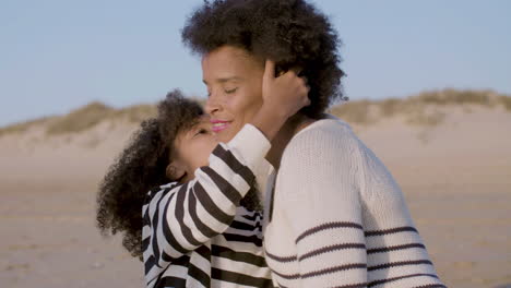 feliz madre e hija abrazándose y besándose mientras se sientan en la playa al atardecer