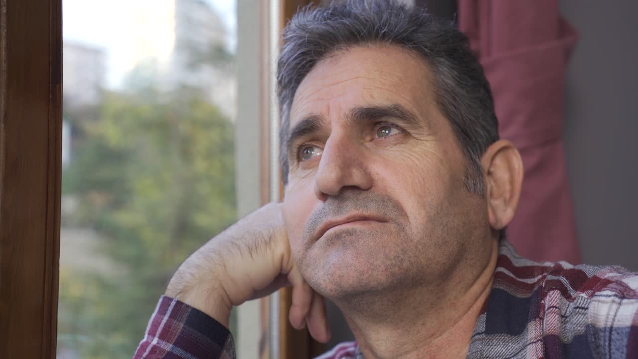 The man is at home watching out the window and is thoughtful.