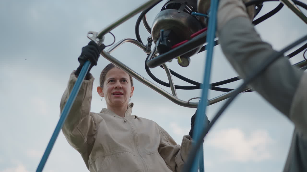 colleague working preflight setup as woman secures burner to hot air balloon frame poles while partner holds support rods under cloudy sky in grassy roadside field coordinating flight preparation