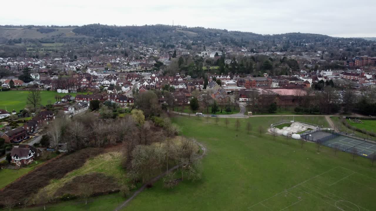 Aerial footage showcasing Reigate town's charming architecture and surrounding greenery from above Reigate Priory Park. Features houses, trees, open fields, and distant hills under an overcast sky.