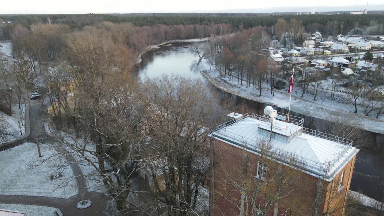 A breathtaking aerial view of Valmiera city, showcasing the winding Gauja River surrounded by snow-covered landscapes and residential areas under a wintry sky.