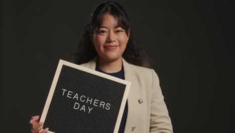 studio portrait of female teacher standing against black background holding up notice board reading teachers day