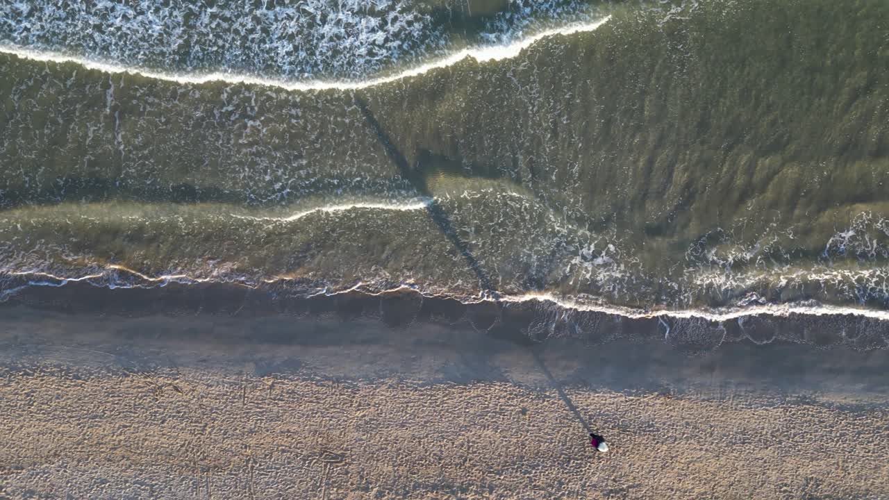 vuelo de dron con vista aérea sobre una playa filmando las olas que terminan en la orilla y una persona caminando con su sombra muy larga por la tarde en la hora dorada en invierno en valencia españa