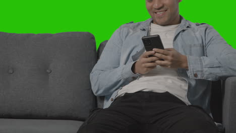 close up of young man sitting on sofa looking at mobile phone against green screen