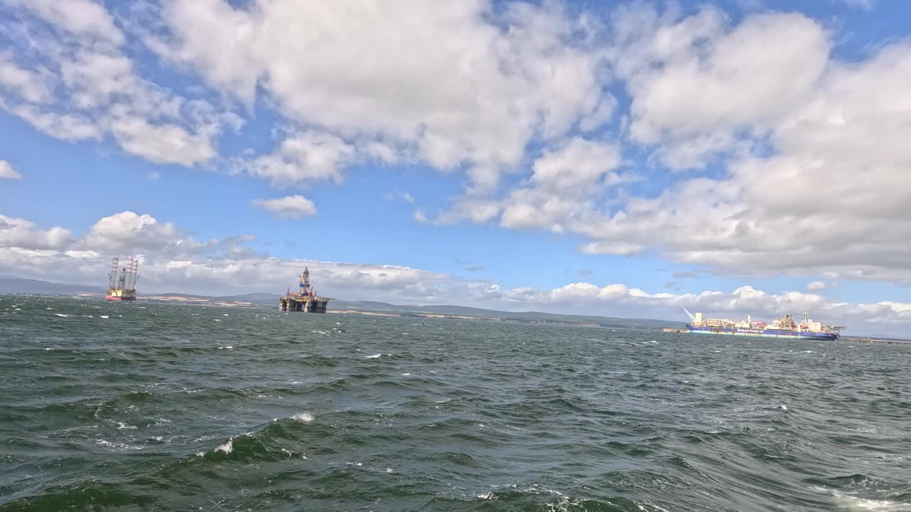 Wide-angle view of rough sea waves, sailboats, and fishing boats under partly cloudy skies in Cromarty, Scotland. Handheld camera pans across dynamic marine scene