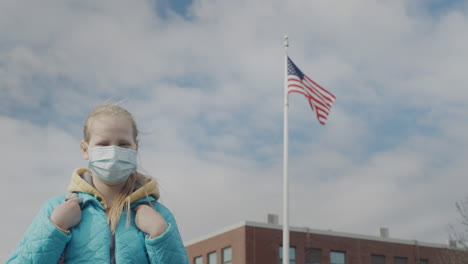 a schoolgirl in a protective mask stands against the background of a school and a us flag.