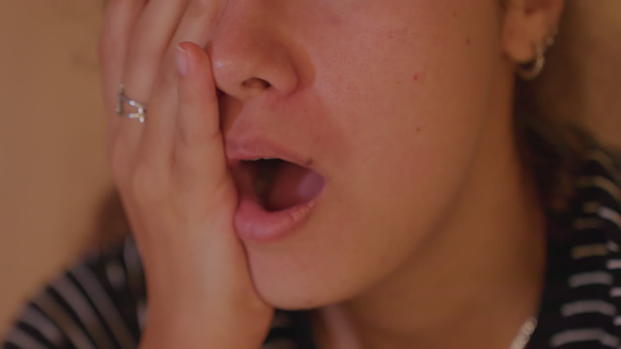 Primo piano di donna che ride e sorpresa, con un dettaglio della camicia a righe. Porta una mano alla bocca, una risata gioiosa si trasforma in un leggero sbadiglio. Luce calda e accogliente, una conversazione rilassata a tarda notte, una reazione da vlog, mento e labbra espressivi.
