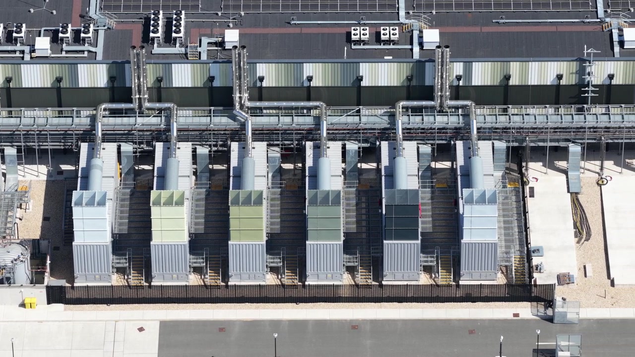 High angle drone view of a massive data center facility featuring HVAC cooling units, power substations, and modern architecture powering cloud networks and AI systems.