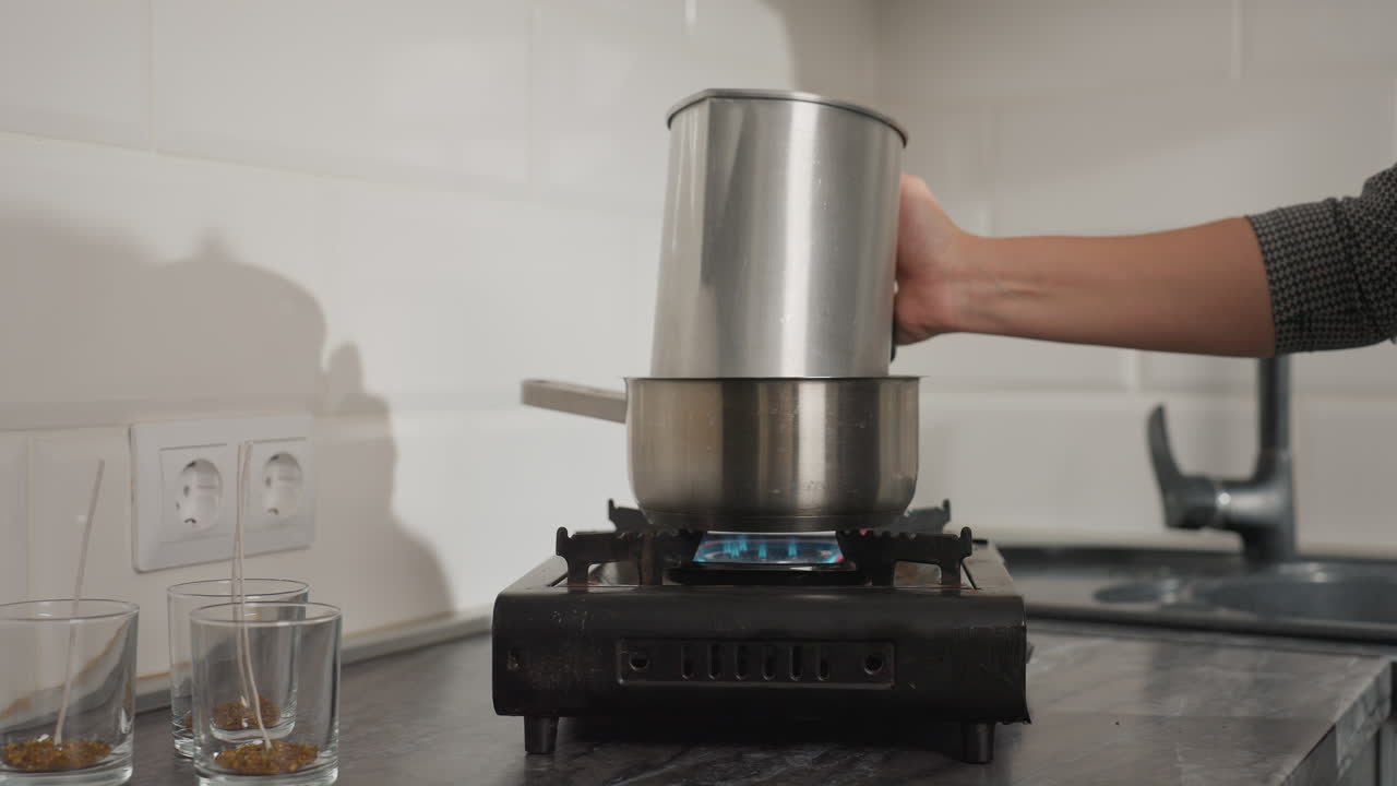 Young woman places stainless jug into pot of water on gas stove, gently releasing jug to float as blue flame burns beneath pot, with glass cups containing wicks placed nearby on kitchen counter