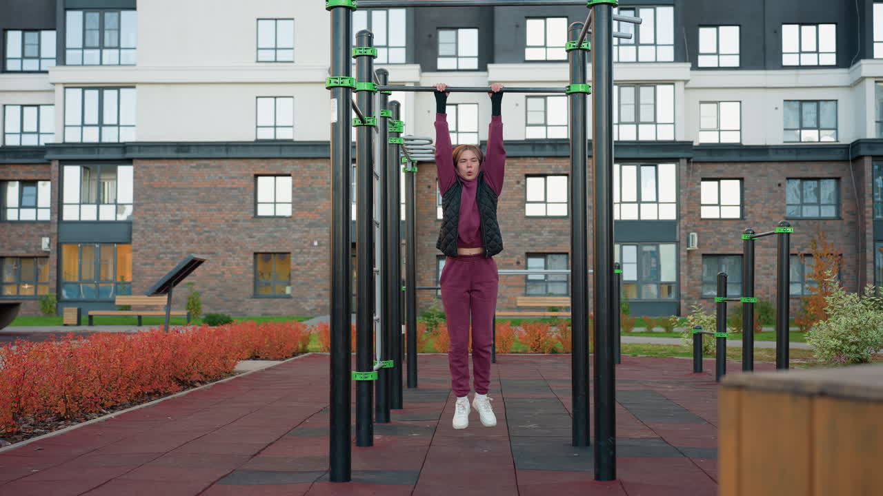 Workout lover swaying while swinging on horizontal pull up bar in urban playground with modern building facade and autumn foliage, showcasing dynamic movement strength and outdoor fitness energy