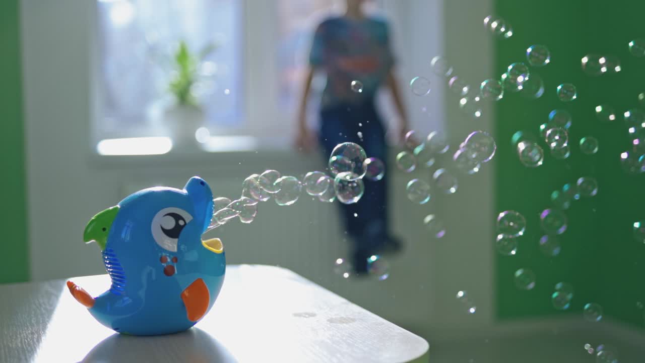 Many small bubbles scatter in one direction from the special appliance on the table indoors. Pretty device in a form of blue fish for creating bubbles on the blur background with a boy.