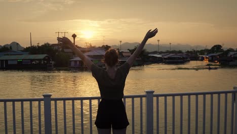 young attractive female tourist standing on harbor whard with raised hands at sunset in thailand