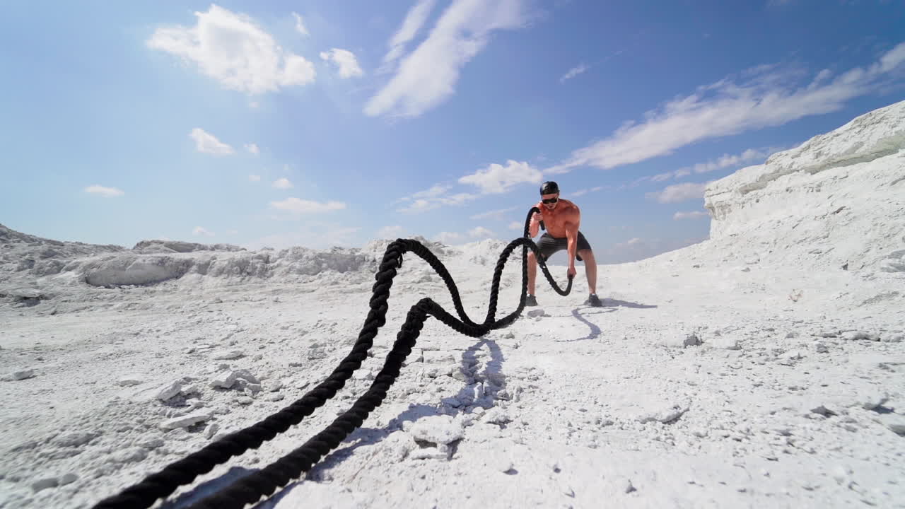 Muscular man with black ropes on white hill under blue sky. Athletic male performs Battle Ropes exercise during an outdoor cross fitness workout. Slow motion.