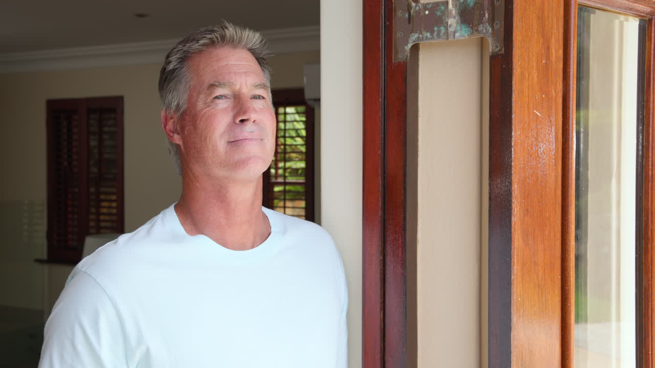 Senior man in white shirt standing by door, looking thoughtfully outside, at home