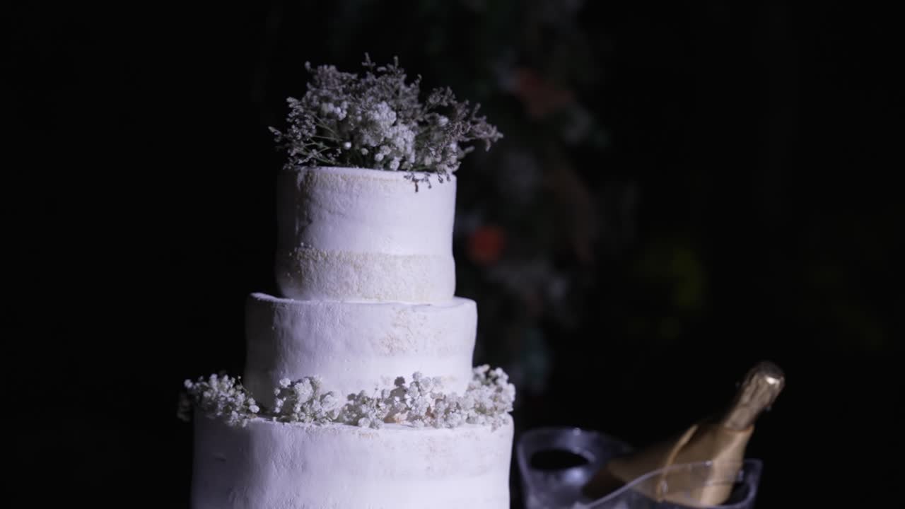 Enchanting white cake crowned with delicate blooms beside bubbly in shadowy elegance