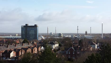 Aerial-descending-view-over-park-trees-to-industrial-townscape-with-blue-skyscraper,-Merseyside,-England