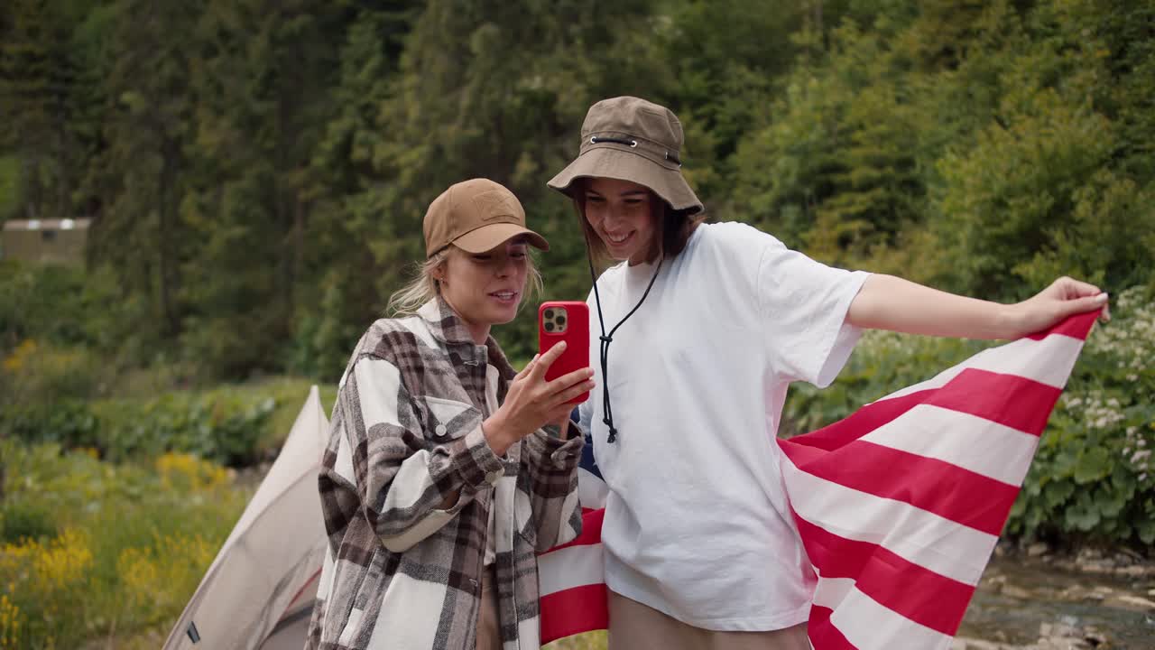 una chica rubia con una gorra muestra a una chica morena con una camiseta blanca con una bandera estadounidense, una foto que fue tomada durante una caminata en el bosque