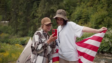 A-blonde-girl-in-a-cap-shows-a-brunette-girl-in-a-white-T-shirt-with-an-American-flag,-a-photo-that-was-taken-during-a-hike-in-the-forest