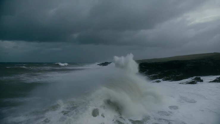 Des vagues océaniques déferlantes se brisant contre un cap rocheux sombre, entraînées par une houle venant de la gauche.
