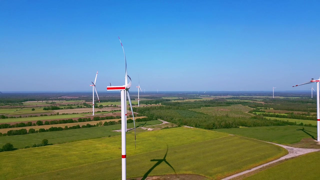 Riprese aeree con drone che sorvolano turbine eoliche rotanti sopra vasti campi agricoli e campagna sotto un cielo azzurro limpido e un paesaggio rurale