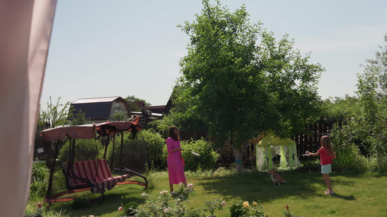Caucasian Mother Coaching Daughter In Backyard Badminton Practice, Racket And Shuttlecock Action, Pink Dress, Focused Practice, Sunny Scene With Fence And Swing, Playful Training Moment